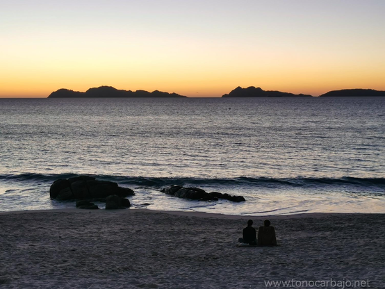 Atardecer, justo después de la caída del sol. En priner plano la arena con la silueta de dos personas sentadas. El agua, unas rocas, el mar  y las islas Cíes al fondo.