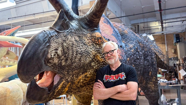 Adam Savage stands with arms crossed next to a life-sized triceratops model in the American Museum of Natural History's workshop.