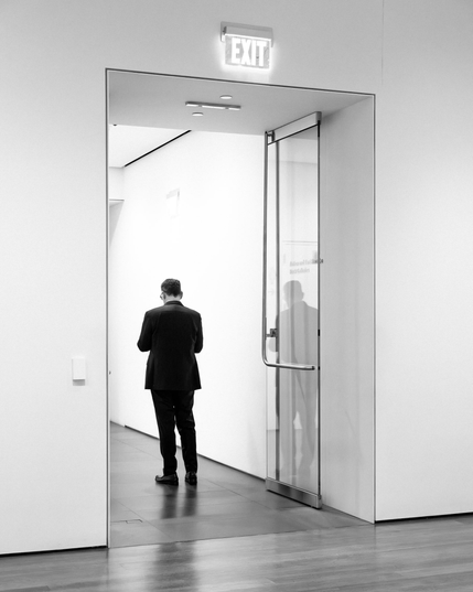 Black and white full length photo of a man in a dark suit facing away from the camera in a doorway. The walls of both the room that the photo is taken from and the room beyond are very nearly white.