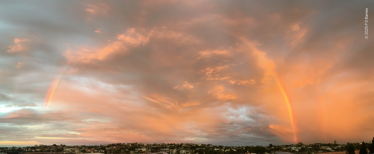 Looking east, the pink light from the setting sun lights up rain clouds drifting across the sky, turning soft blankets of rain into diffuse clouds of pink, and sketching the outlines of a rainbow, marking in glowing colour where the rain is heavier.
