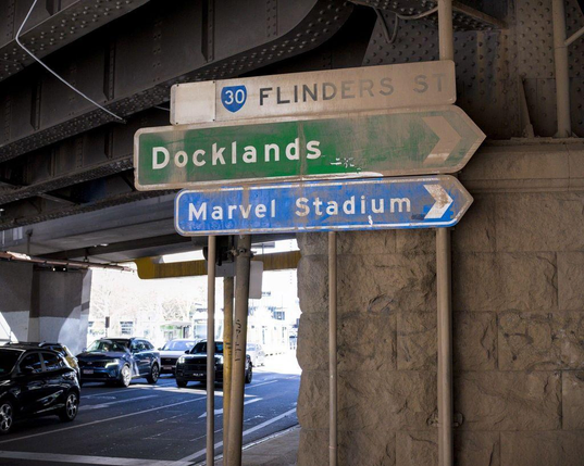 Photo showing three large road signs erected on metal poles. The signs are next to a massive concrete pillar with a stone facade. This pillar is one of several that are holding up a series of elevated train lines that cross over the roads that run beneath. The signs read, from top to bottom, "Flinders St" (the cross street at this intersection), "Docklands" (the suburb to the right of this intersection), and "Marvel Stadium" (the point of interest to the right of this intersection).