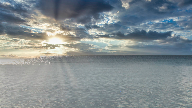 Ein Blick über eine ruhige Ostsee zum Horizont. Dort steht die aufgehende Sonne die mit ihren Sionnenstrahlen die Wolken am Himmel durchbricht.