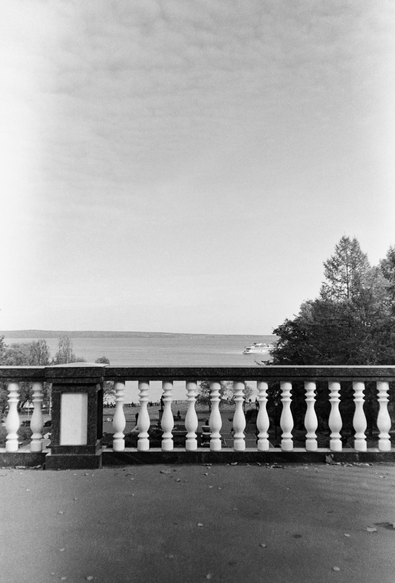 The black-and-white photo shows a view of Lake Onega from the terrace. In the center of the frame is a white fence, behind which is a view of the water surface. In the background, there are trees and a small ship on the horizon.