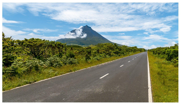 Eine schnurgerade Straße führt über ein von niedrigen Pflanzen bewachsenes Hochplateau. Im Hintergrund ragt ein Vulkan hervor.
