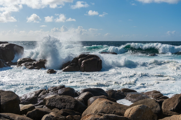 Waves crash against rocky shores under a blue sky with some clouds. The sea is moving energetically, creating white foam as it hits the rocks.