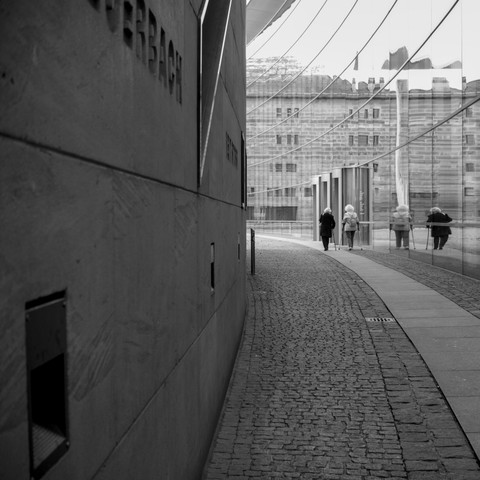 The black-and-white photo shows a narrow, cobblestone walkway curving between two large, modern buildings. On the left, there is a wall with engraved or inlaid lettering. On the right, a glass facade reflects the scene and the buildings opposite. In the background, two people are visible walking along the path. Both individuals are clearly reflected in the glass facade.