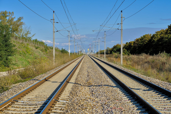 Das Foto zeigt eine gerade Eisenbahnstrecke mit zwei Gleisen, die sich in der Ferne perspektivisch treffen. Links und rechts der Gleise sind grüne Büsche, Bäume und Gräser zu sehen. Mehrere Strommasten und Oberleitungen verlaufen entlang der Strecke. Der Himmel ist klar und blau, mit nur wenigen Wolken am Horizont. Es handelt sich um eine typische Landschaftsaufnahme eines modernen Bahnabschnitts.
