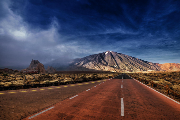 Lange, dunkle Strasse mit rötlichem Asphalt führt direkt auf einen grossen, kahlen Berg zu. Himmel tiefblau mit dramatischen, dunklen Wolken. Die Landschaft ist trocken und buschig, links ein Felsvorsprung mit einem kleinen braunen Wegweiser.