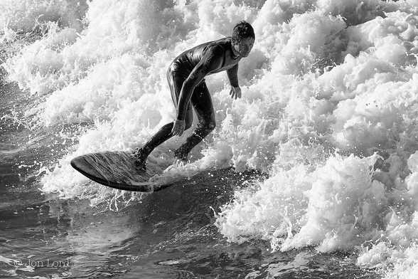 This is a black and white photo in landscape format of a man surfing ahead of a breaking wave. Hermosa Beach, California (2014).

From the lower right corner running diagonally across the image to the upper left corner is a mass of brilliant white foaming surf. In the lower corner is the dark and fairly smooth water ahead of the breaking wave. In the centre of the photo is our male surfer, he is wearing a one-piece black wetsuit. The surfboard is black and heading towards and to the left of the camera. Our surfer is standing, slightly crouched, at right angles to the board and is facing to the camera's right, his arms outstretched as he maintains balance. The front of the board is visible with a thin stream of water running over it's surface, the back half of the board is hidden inside the deluge of water from the surf which is about the height of the surfer. 