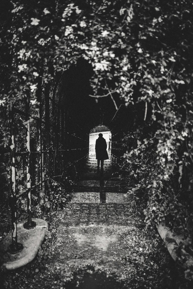 The image presents a monochrome, atmospheric scene of a person standing at the entrance of a dark tunnel or archway. The composition is centered, drawing the eye towards the figure at the tunnel's opening.

The tunnel itself is the dominant element, framed by lush vegetation that arches overhead and along the sides. The foliage is dense, with visible leaves and branches creating a natural frame and adding texture to the image. The tunnel's interior is dark, contrasting sharply with the brighter light at the exit.