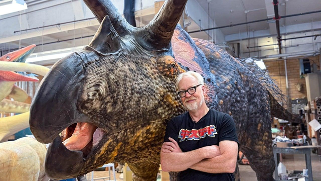 Adam Savage stands with arms crossed next to a life-sized triceratops model in the American Museum of Natural History's workshop.