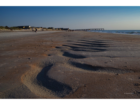 Sandy beach at sunrise with ripples in the foreground, people walking in the distance, calm sea, and a pier extending into the ocean under a clear sky.