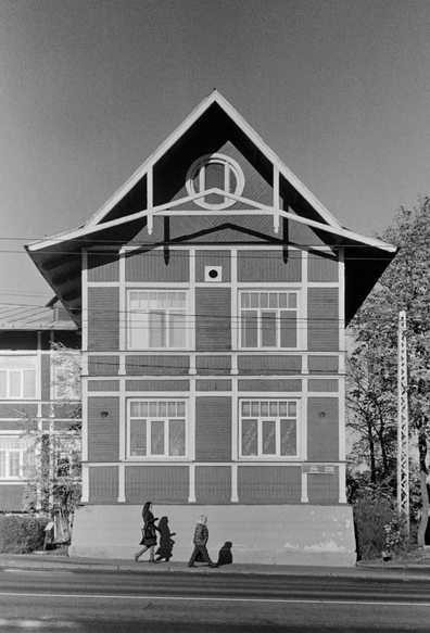 This black and white photo features a building of old Finnish architecture, with a young and an elderly woman walking past. Their shadows are cast upon the wall, and a road stretches out in the foreground, adding to the vintage atmosphere.