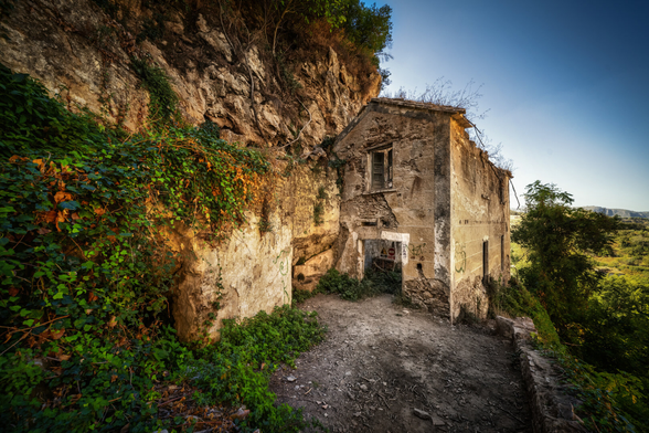 Altes, baufälliges Steingebäude klebt an einer steilen, bewachsenen Felswand. Üppiges, grünes und teilweise vergilbtes Efeu überwuchert die linke Wand. Vor dem Haus liegt ein unbefestigter, schmutziger Weg, der nach rechts hin abfällt und einen Blick auf eine sonnige, grüne Landschaft und einen klaren, blauen Himmel freigibt. Das Licht fällt stark auf die Szene, was die Textur der verwitterten Steine hervorhebt.