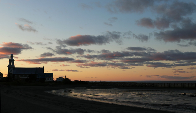 Photograph of a sunset over a calm cove, with a Catholic church and its cemetery to the left of the image, and a stone pier extending into the water. The sky is blue at the top, fading to shades of yellow, orange, and pink towards the horizon, with a few grey clouds reflecting pink. The calm water of the cove mirrors the blue of the sky.

Photographie d'un coucher de soleil sur une anse de mer calme, avec une église catholique et son cimetière à gauche de l'image, et un quai en pierre qui s'avance dans l'eau. Le ciel est bleu tout en haut et se dégrade en teintes de jaune, orange et rose vers l'horizon, avec quelques nuages gris où se reflète du rose. L'eau calme de l'anse reflète le bleu du ciel.