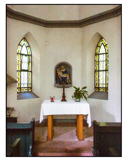 interior of a small chapel, including two windows and the altar, which is basically a wooden table with a candle, a crucifix and a plant on it.