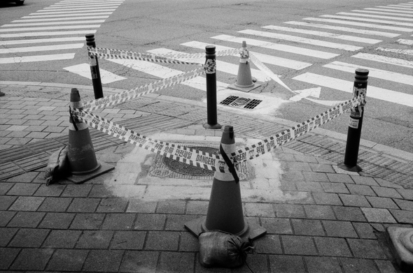 건널목 횡단보도 앞 인도의 맨홀 뚜껑을 중심으로 볼다르와 러버콘, 안전제일 경계끈을 이용해 출입을 막고 있다. 무언가 시공을 한 뒤 굳히는 모양이다.