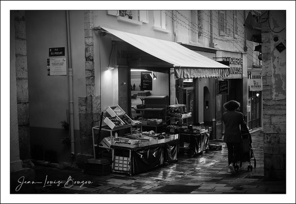 This black-and-white photograph portrays a quiet morning scene on a narrow street, likely in a European town judging by the architectural style and street layout. The wet cobblestone pavement reflects the light, suggesting it has recently rained, adding a glistening texture that enhances the mood of the image.

The focal point is a small shop or market stall under a striped awning, with an assortment of crates and baskets filled with goods—possibly fruits, vegetables, or other produce—carefully arranged along the wall. The lighting from the shop casts a soft, inviting glow onto the street, contrasting beautifully with the darker surroundings.

On the right, a person dressed in a coat and hat, seen from behind, pushes a baby stroller. This figure adds a sense of daily life and quiet routine, emphasizing a moment of solitude or simplicity within an urban environment.

The texture of the old buildings, the wet street, and the subtle light interplay create a nostalgic and timeless atmosphere. Black-and-white photography here emphasizes shape, light, and shadow rather than color, inviting the viewer to focus on the composition and the emotions evoked by the scene.
