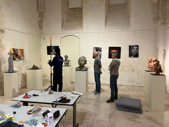 Photographie d'une exposition en cours d'installation. Au premier plan, 2 tables couvertes d'outils. Au milieu, 3 personnes (2 hommes et 1femme) regardent vers la gauche pour régler l'alignement d'une photographie sur le mur. Autour d'eux, 5 sculptures sont posées sur des socles blancs et 5 photographie sont accrochées au mur de manière bancale.