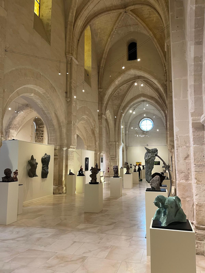 Photographie de l'intérieur d'une église gothique servant de salle d'exposition. La photo est prise en contre plongée et on voit les voûtes de pierre très hautes et les éclairages qui font une sorte de constellation au dessus des sculptures posées sur des socles blancs ou accrochées au mur.