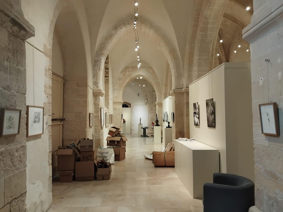 Photographie d'un couloir dans une église gothique servant de salle d'exposition. On y voit des photographies et des dessins dans des cadres accrochés aux murs de part et d'autre. Des cartons d'emballage jonchent le sol.