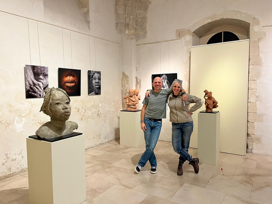 Un homme et une femme en jeans sont au centre de la photo, debout, souriant et se tenant par l'épaule. Ils sont entourés de 3 sculptures et de 4 photographies dans une salle dallée de pierres aux murs blancs