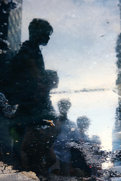 Through a puddle in the asphalt there is a silhouette of a young child with medium length hair. The outline of their face is sharp enough to make out. Beyond them are three other silhouettes behind them, blurry and obscured by the surface of the puddle.