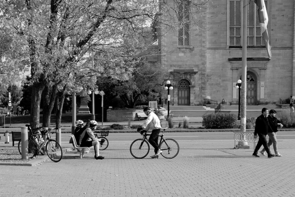 On the street outside the National Gallery. Across the centre of the image, running left to right, is a perfect line of bikes, riders sitting on a bench, another rider astride his bicycle, and two pedestrians walking toward the left edge of frame. Behind them are a big leafy tree at left and the facade of the church across the street at right. 