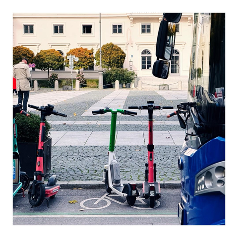 Von rechts kommt ein blauer Bus ins Bild, auf der gegenüberliegenden Straßenseite stehen E-Rollen in verschiedenen Farben 