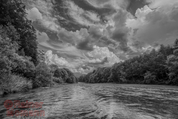 Schwarzweißfotografie im Format 3:2 Landscape.
Der Fluss Vils, an einer breiten Stelle und viel Wasser führend, welches schnell fließt. Beide Ufer mit dichtem Wald gesäumt. Darüber durchscheint die Sonne einen durchwachsenen Wolkenhimmel, welcher immer wieder einen Regenschauer, zwischen den sonnigen Abschnitten bringen kann.