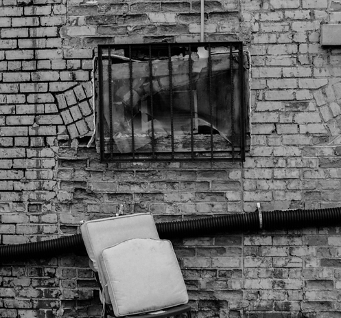 A black and white photo of a very low to the ground barred window in a brick wall.  There is corrugated plastic conduit running along the wall underneath and seat cushions stacked on a barely visible chair.