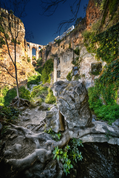Ein schmaler, verwinkelter Pfad schlängelt sich zwischen Felsen und üppigem Grün hindurch. Beidseitig ragen steile Felswände empor, rechts sind alte, verlassene Gebäude mit zerfallenen Fenstern eingebettet. Über dem Pfad wachsen Wurzeln und Pflanzen, die sich um die Steine ranken. Im Hintergrund spannt sich eine hohe Steinbrücke über die Schlucht, während der Himmel tiefblau und klar ist.