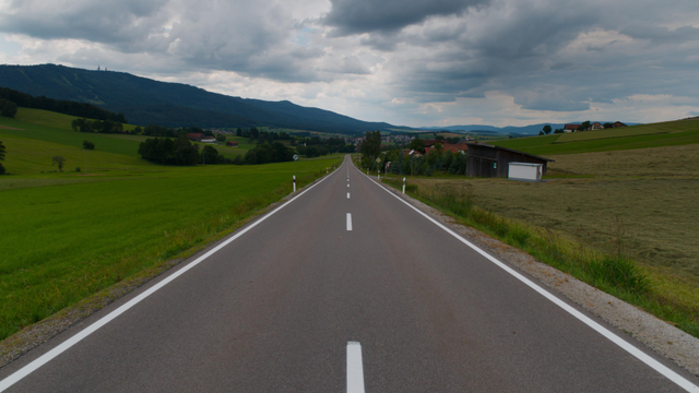 A straight, asphalted road goes from the whole bottom of the picture almost up to the horizon. To the left and right, there are green and yellow fields and a few small buildings. At the far end, there is a village and a large mountain. Above the scene are some dark clouds.