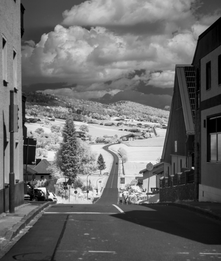 Monochrome picture with an outer-world effect caused by filming in infrared. A road starting in a hill-top village, with houses left and right, meanders away from the camera, visible for quite some kilometers. To its left and right are very bright shining fields, almost white, with a few trees. In the background, there are other hills and large clouds.