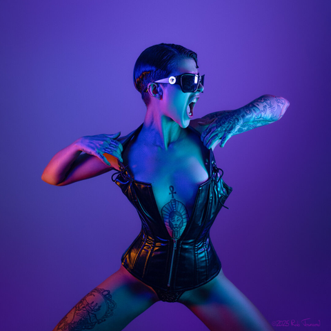 A photo of art model Sofia Predrazzoli by photographer Rab Townsend. The model is a woman with short, slicked back, dark hair. She is posing against a violet background, wearing a leather corset, that zips only halfway up, revealing a tattoo on her sternum. She is wearing sunglasses, and her face is turned to the right of frame, with her mouth open as if mid-shout. She is pulling the edges of the corset open with each hand, with elbows extended outward to her right and left. Her arms and hands are covered with tattoos, as is her right thigh. Her skin is lit with a combination of blue, cyan, and orange light. She is wearing lacey black underwear, peeking out beneath the corset. The image evokes a fun rockstar energy on a fashion shoot.