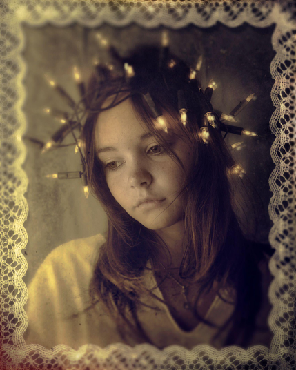 Portrait d'une jeune fille au long cheveux bruns, elle porte une chemise blanche et est photographiée à hauteur du sternum. Elle semble pensive, ses cheveux sont cerclés d'une guirlande composées de bougies électriques allumées. Les couleurs sont sépias/jaunes