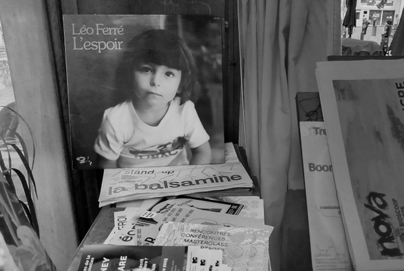 Photo noir et blanc.
À gauche, la pochette d'un 45 tours de Léo Ferré, l'espoir.
Image d'un enfant nous regarde.
Devant cette pochette, des prospectus culturels.
Sur la droite, rideau et un petit carré de fenêtre avec vue sur l'extérieur.
À droite, journal du @nova

