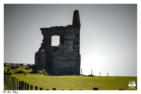 A dramatic photograph, taken near Stanley, Tasmania, showing the stone ruins of a chimney and wall silhouetted against a bright, near white sky.

The dark, crumbling structure stands on a grassy hill, creating a strong contrast.

A wooden post-and-wire fence runs along the foreground. Signed Kev Peirce.