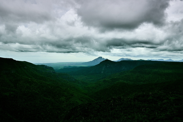 A view of lush, green hills extending towards the ocean under a cloudy sky. Distant mountain peaks create depth in the landscape.