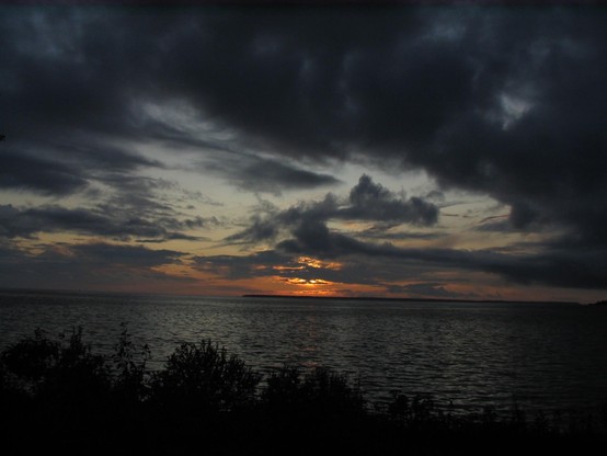 Sun setting in the distance over water with dark clouds in the sky.