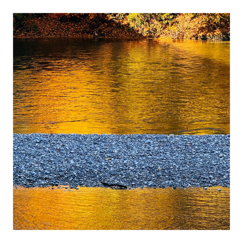 Im Wasser eines Flusses spiegelt sich das orangefarbene Laub. Quer durchs Bild geht ein Streifen mit grauem Kies.