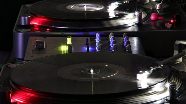 The image shows a close-up, 30-second exposure of active vinyl turntables and a mixer.

Both turntables are illuminated by their various onboard lights, although the one further back is also illuminated by lights on the mixer. The needle cartridges are blurred from motion, and are reflected in the surfaces of the records being played.
  
Between the turntables, there's a DJ mixer with various knobs and buttons. Some of these are illuminated by the blue power light, as well as the green lights of the volume meter, adding to the colorful lighting effect.

The equipment has been set up on a table in the dark, where the depth of field completely obscures any surrounding detail, which makes the bright lights from the turntables and mixer stand out more prominently.