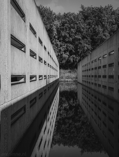 The image is dominated by two massive, concrete structures. Two parallel, tall structures of raw, gray concrete dominate the frame, creating a deep, vanishing perspective. 

A narrow channel of still water separates the two structures, acting as a flawless, dark mirror. This reflective surface captures the perfect mirror image of the walls and windows, creating a powerful sense of symmetry and a strong vanishing point in the center. 

The water's surface is deep black and slightly rippled, providing a rich, inky contrast to the lighter concrete.