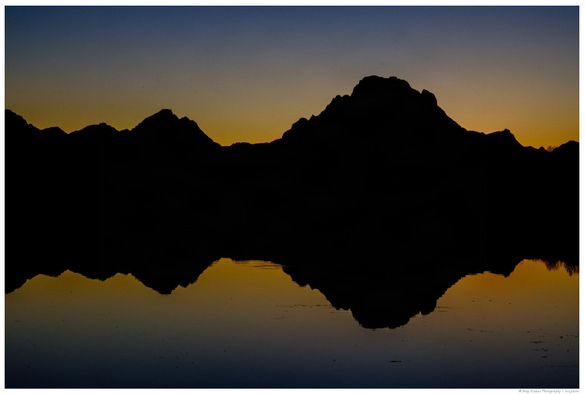 A silhouetted mountain range is reflected in calm water below, with a gradient sky transitioning from deep blue at the top to golden yellow along the horizon, suggesting sunrise or sunset.