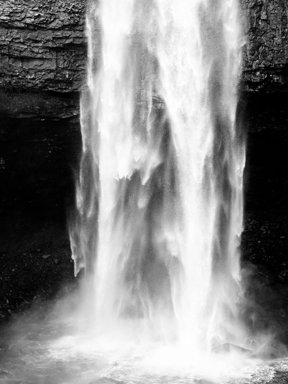 A vertical, black-and-white photograph of a large, powerful waterfall. The image is dominated by the brilliant white, rushing water that falls from the top edge straight down into a turbulent pool at the bottom. The falling water is so forceful that it appears as a thick, nearly solid curtain, with spray and mist billowing out, creating a soft, ethereal texture. The waterfall is framed by dark rock walls on either side. These cliffs provide a sharp contrast to the bright white water. The texture of the cliff face at the top is rough and layered, suggesting deep, natural stone formations. The overall impression is one of immense power, movement, and high contrast, emphasizing the force and beauty of the natural element against the stark, dark rock.