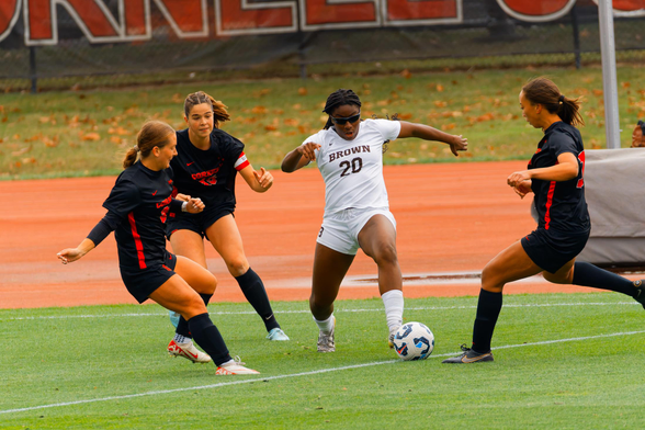 A black women wearing a white shirt with the words BROWN 20 runs forward with the ball while three girls in black jersey and socks with red letters cnverge on her.