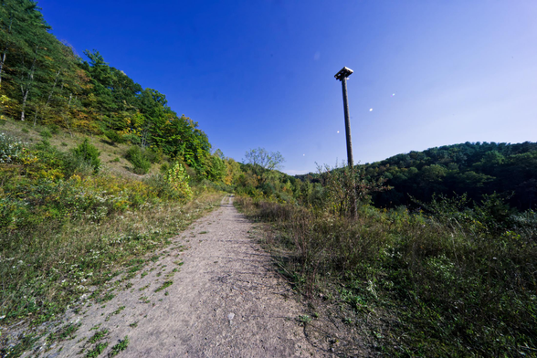 A sandy path runs from the bottom left to the center,  there is a slope with the high end and the low on the left end and low to the right,  a distant hill can be seen to the right,  and a telephone pole with a large box on the top is just a bit right of the path.