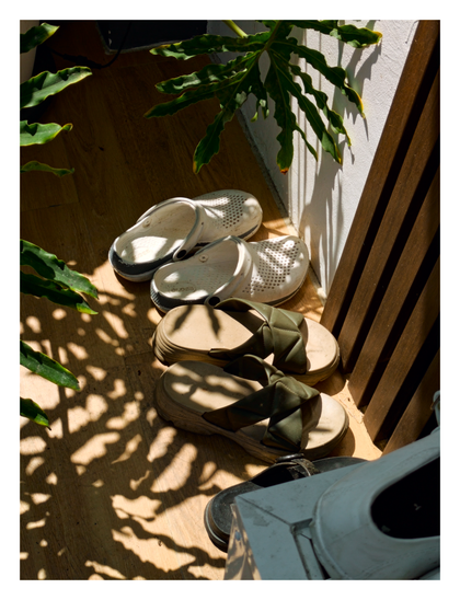 Two pairs of shoes — white crocs and green platform sandals — resting on a wooden floor in a sunlit corner. The scene is covered in a dappled pattern of light and shadow cast by the leaves of an overhanging plant.