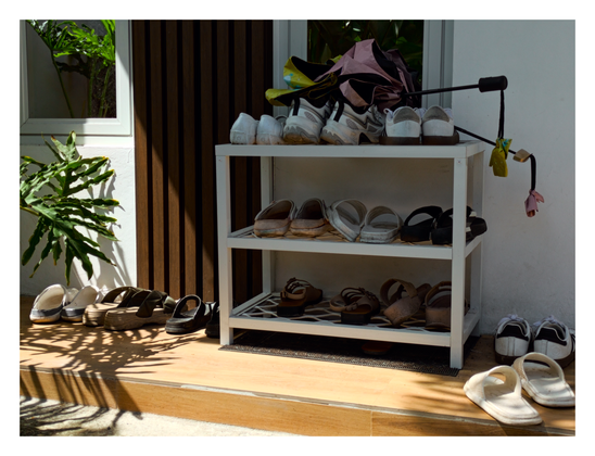 A white, three-tiered shoe rack stands on a wooden-tiled step at the entrance of a home. The rack is filled with various shoes, including sneakers on the top shelf alongside a folded umbrella, and sandals on the lower shelves. To the left, a large, leafy green plant casts distinct shadows across the sunlit ground and step. The background consists of a white wall and a dark brown, vertically slatted door. Additional pairs of shoes are placed on the step around the rack.