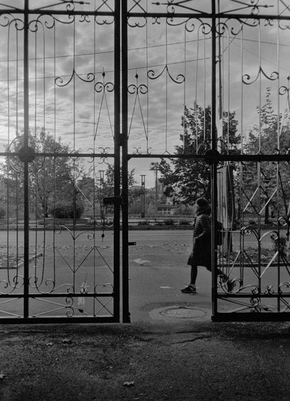 The black-and-white photo shows a high metal gate with a door. Through the gate, you can see a street with a person walking on it.